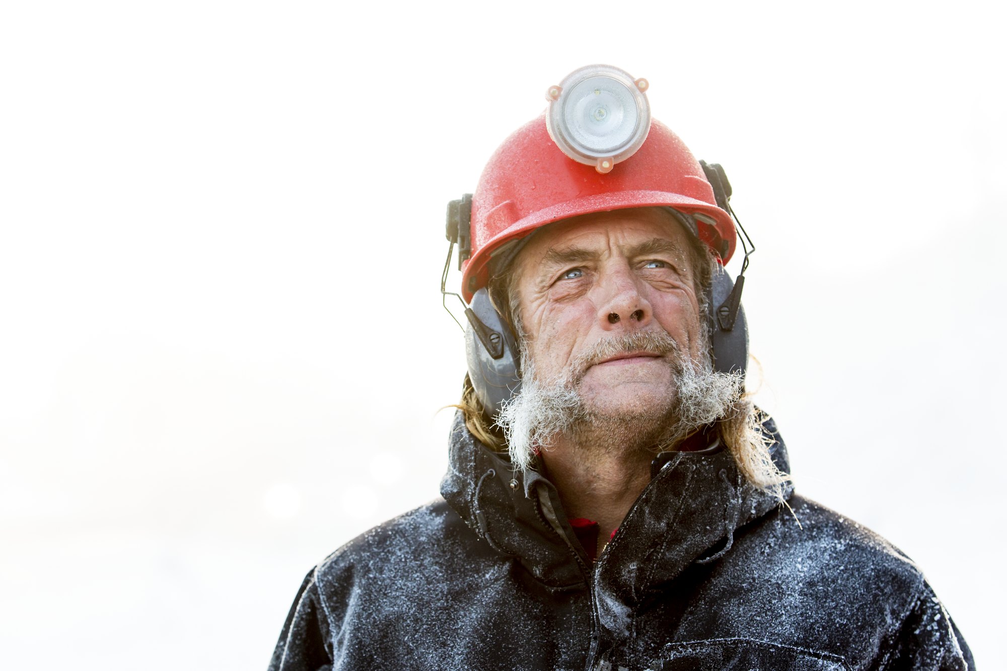 Portrait of a Killington snowmaking crew member at dawn — frost in his beard, red hard hat and headlamp, looking up into the light